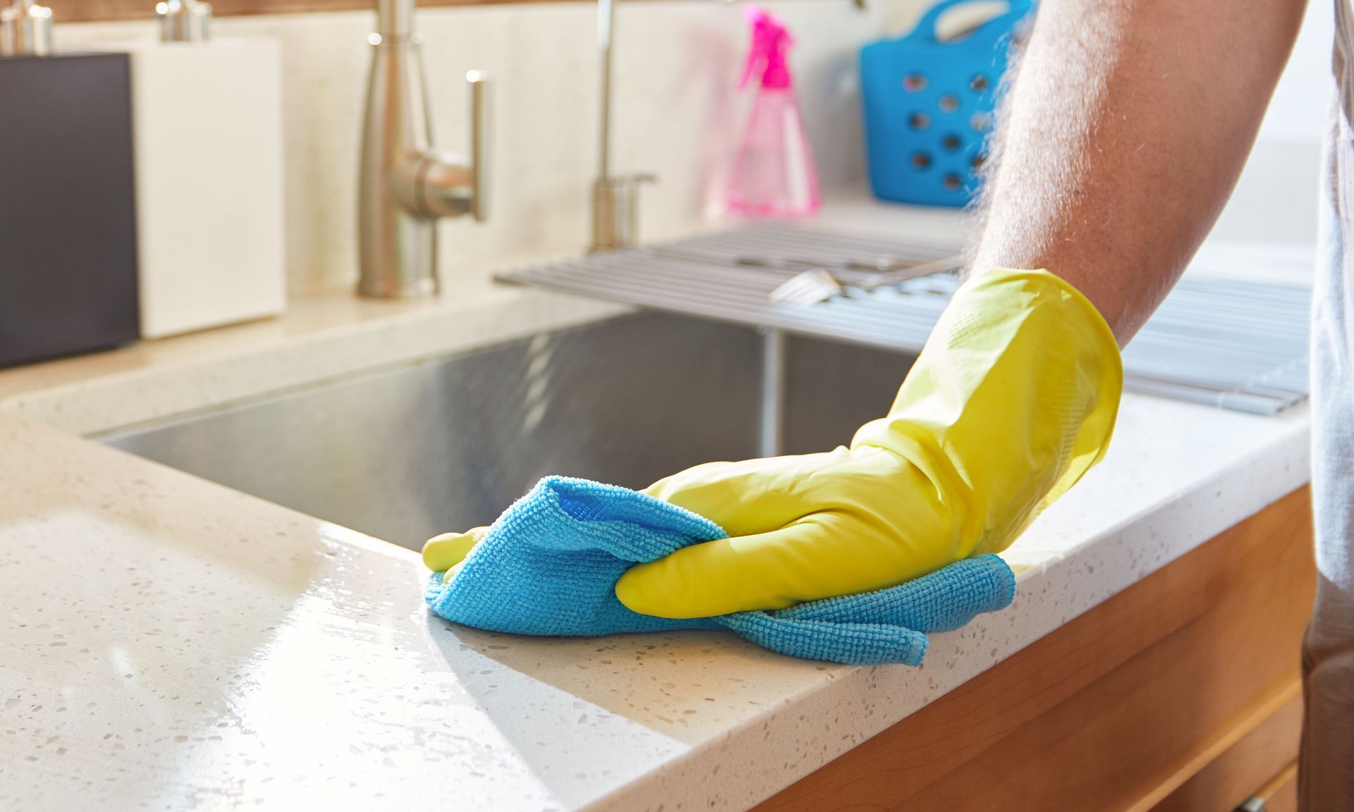 Yellow-gloved hand wiping a kitchen countertop with a blue cloth beside a sink
