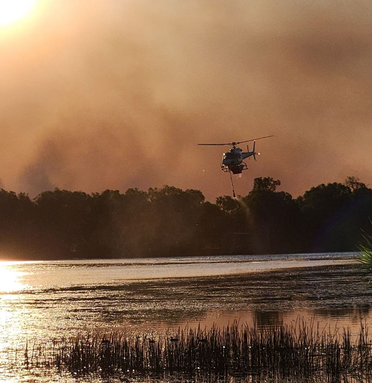 Helicopter dropping water on a fire over a body of water with smoke in the sky during sunset.