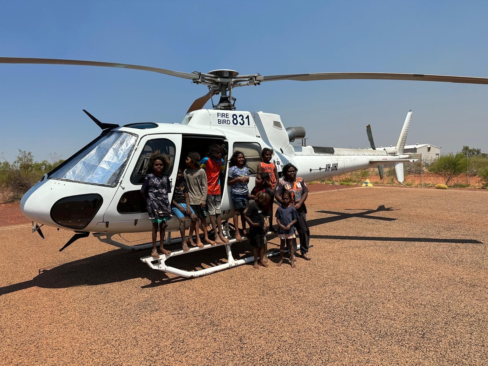 People standing in front of a white helicopter on a gravel area under a clear blue sky.