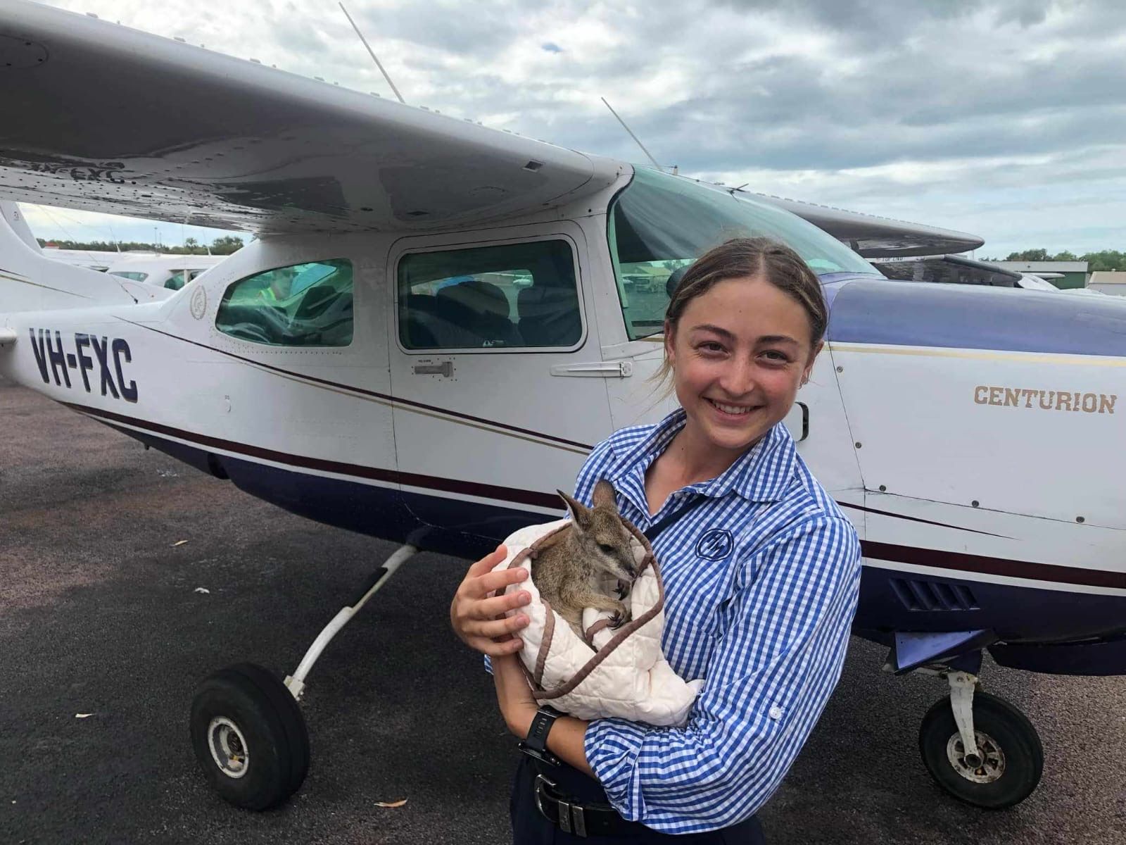 Woman holding a joey in front of a small airplane, smiling. Outdoors, blue and white plane.