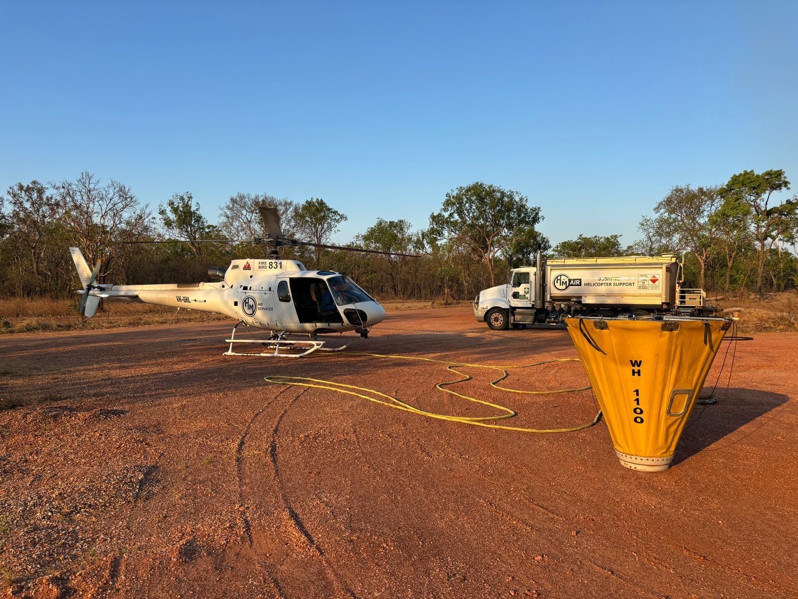 Helicopter with open door and truck on dirt, near a yellow water bucket, in a clearing.