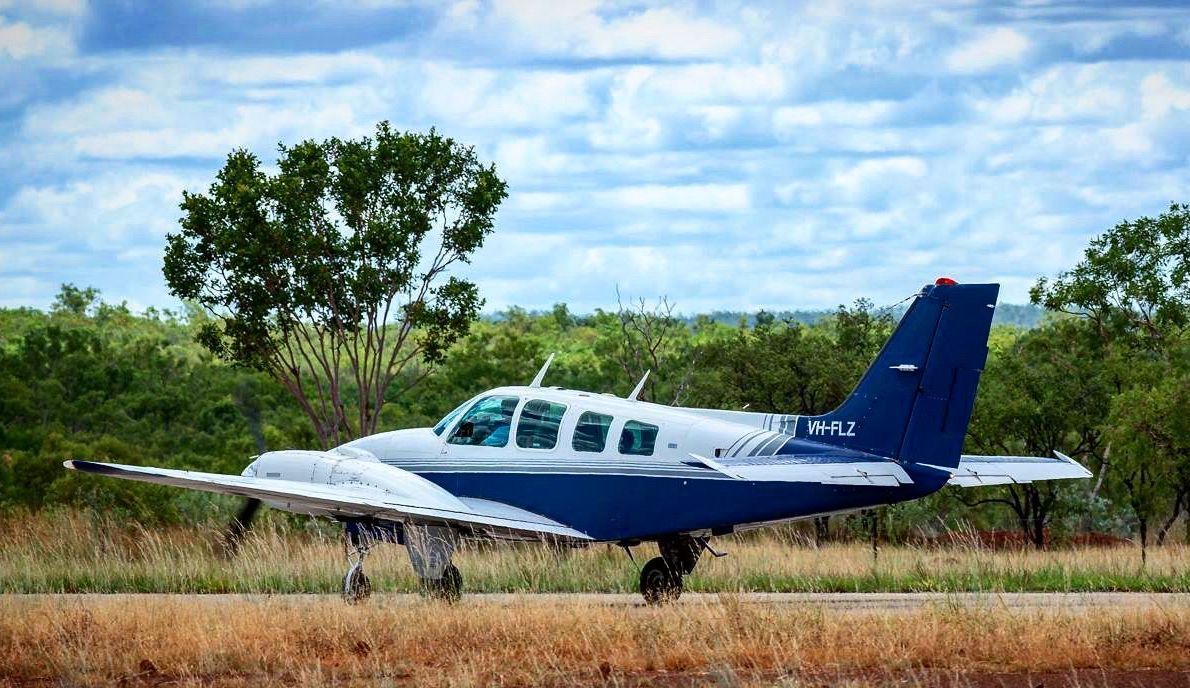 A small blue and white airplane on a grass runway, trees in the background.