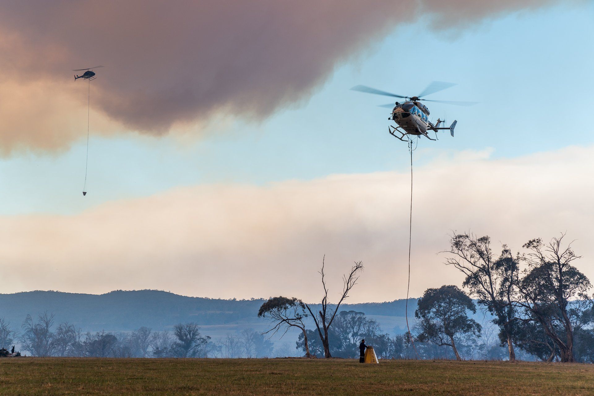 Helicopters dropping water on a wildfire; smoke in sky over a dry field and trees.