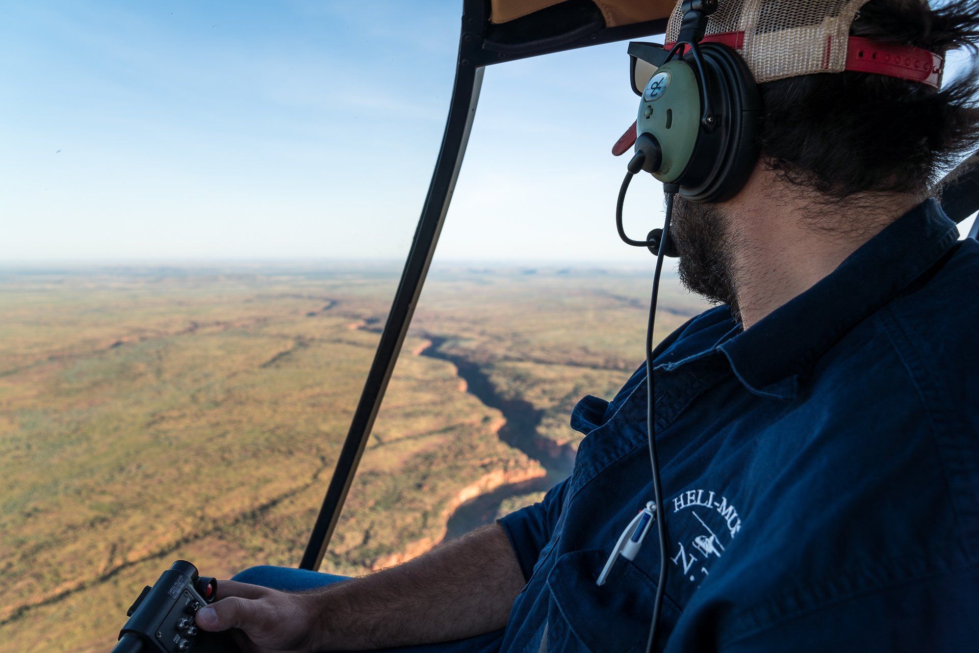 Pilot in a helicopter, looking out over a canyon landscape. Wearing a headset and holding controls.