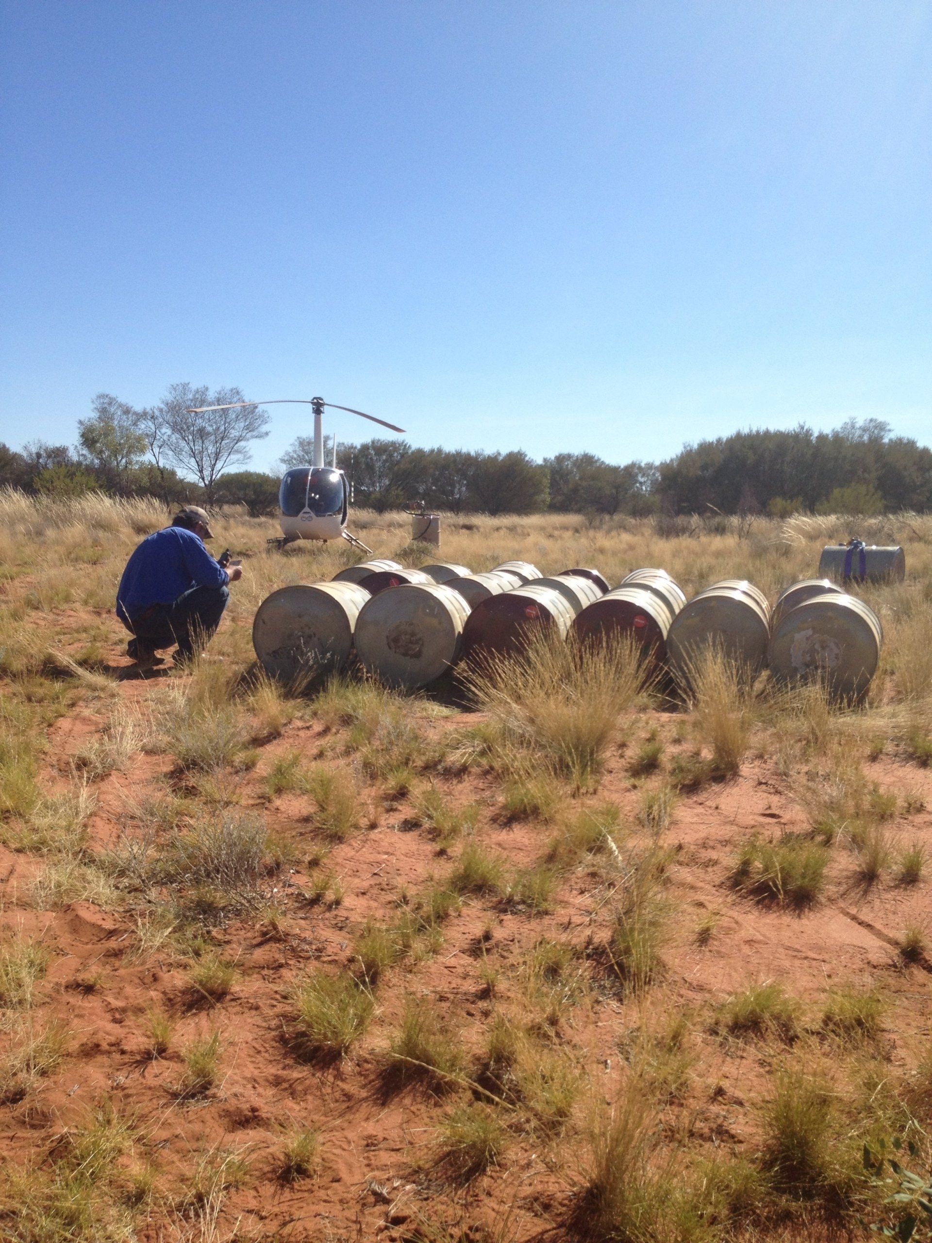Person crouches near barrels on a field with a helicopter in the background under a clear sky.
