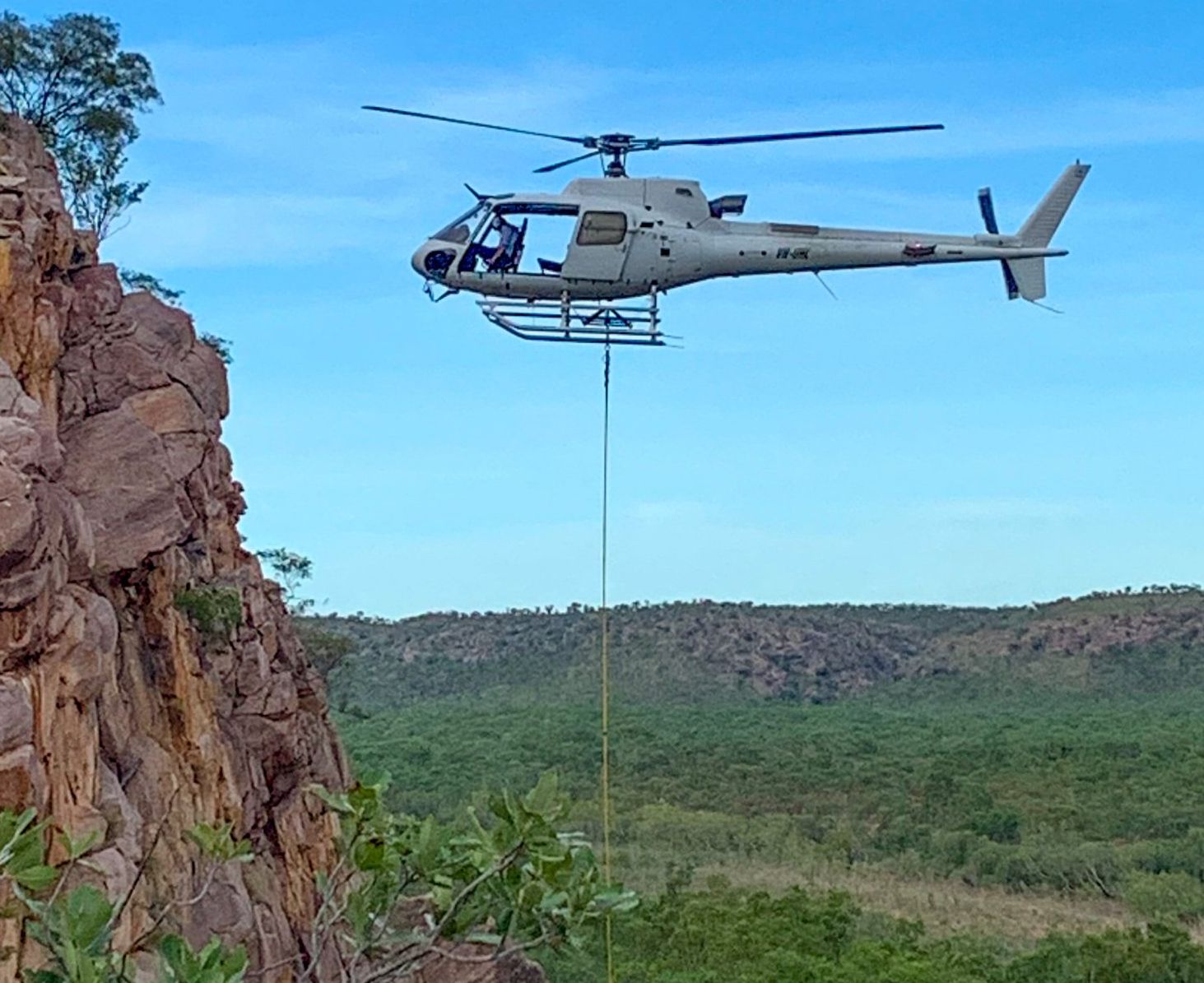 Helicopter flies past a rocky cliff, carrying a long line; green landscape and blue sky in the background.