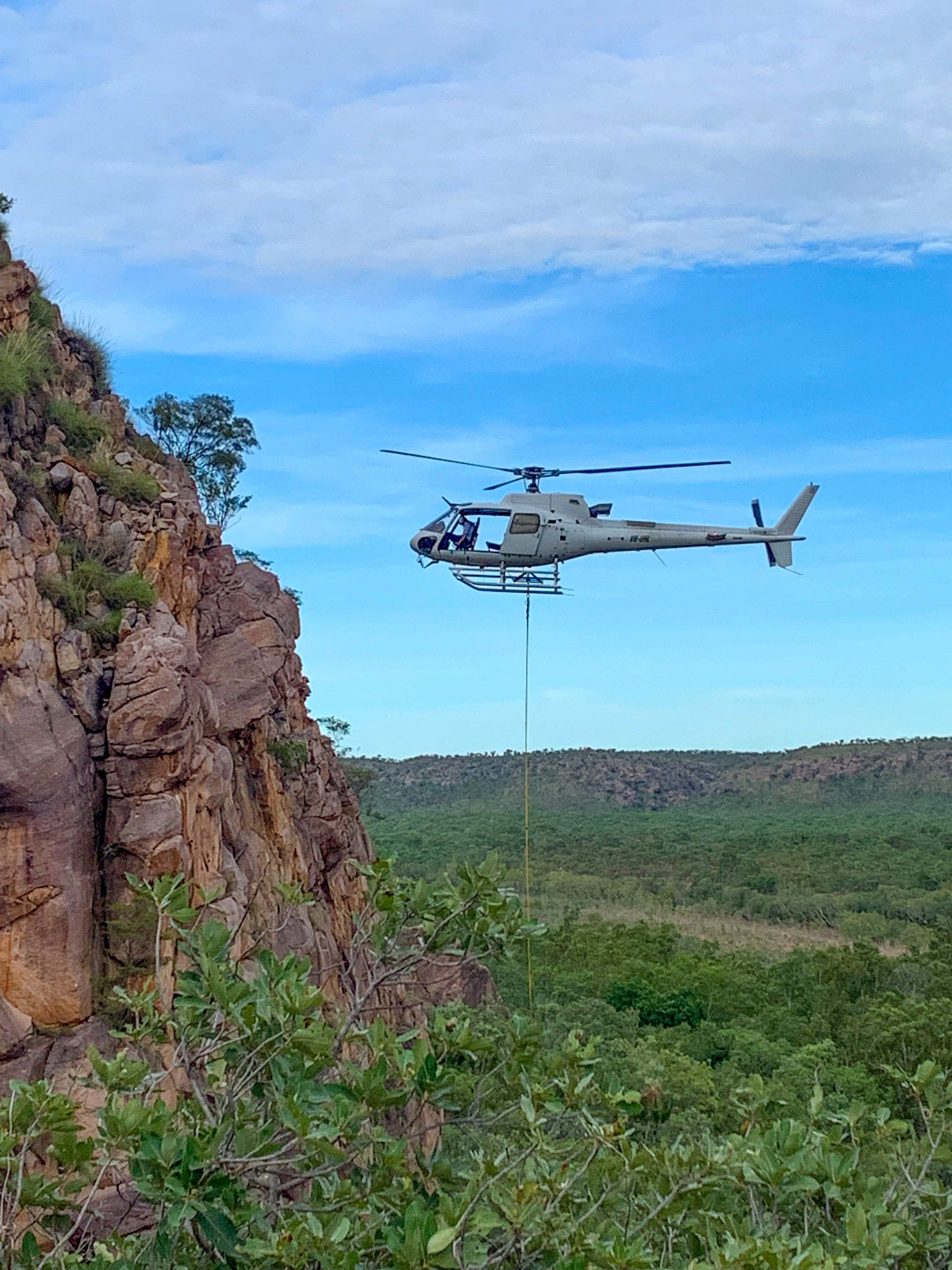 A military helicopter flies over rocky terrain under a blue sky.