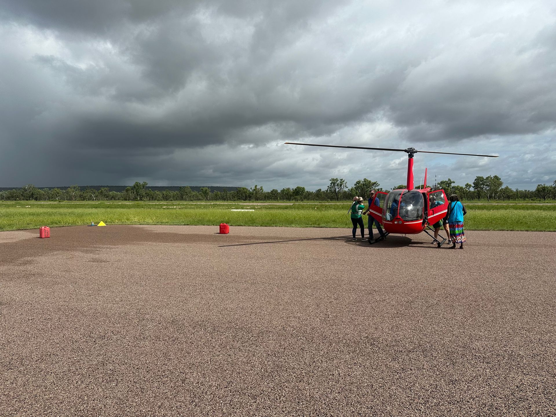 Red helicopter on a gravel landing pad with people and a stormy sky overhead.