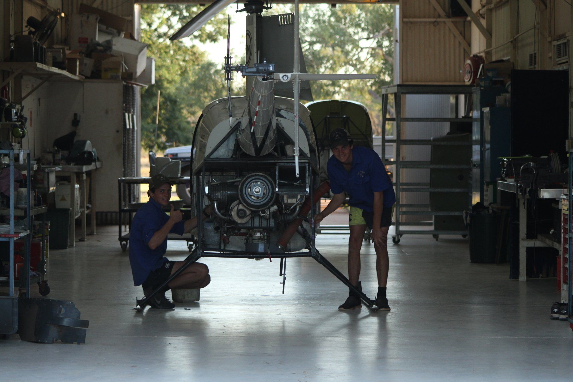 Two people working on a helicopter inside a workshop. One kneels, the other stands, both in blue shirts.