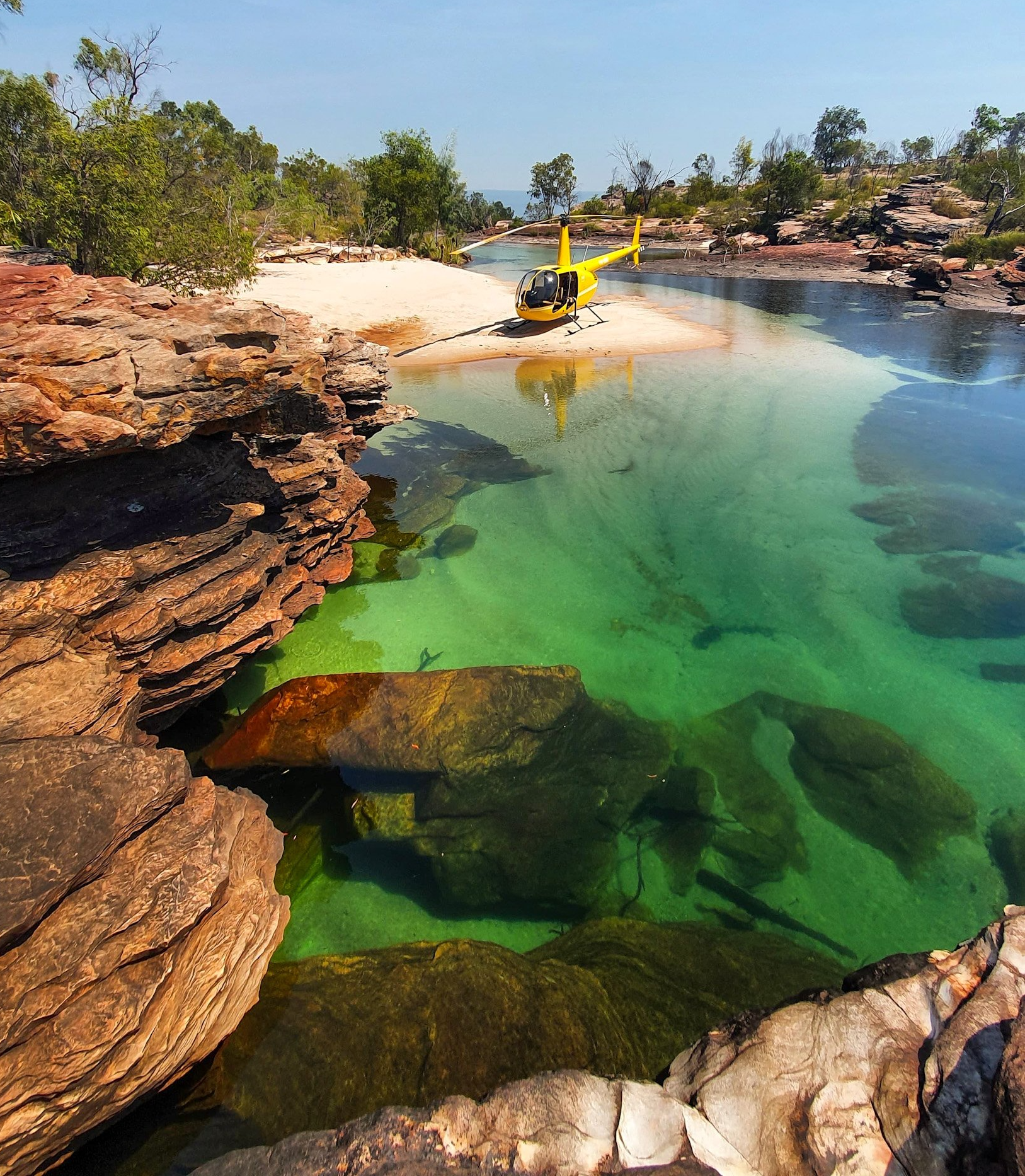Yellow helicopter parked on a sandy bank next to a clear turquoise waterhole. Rocky landscape.
