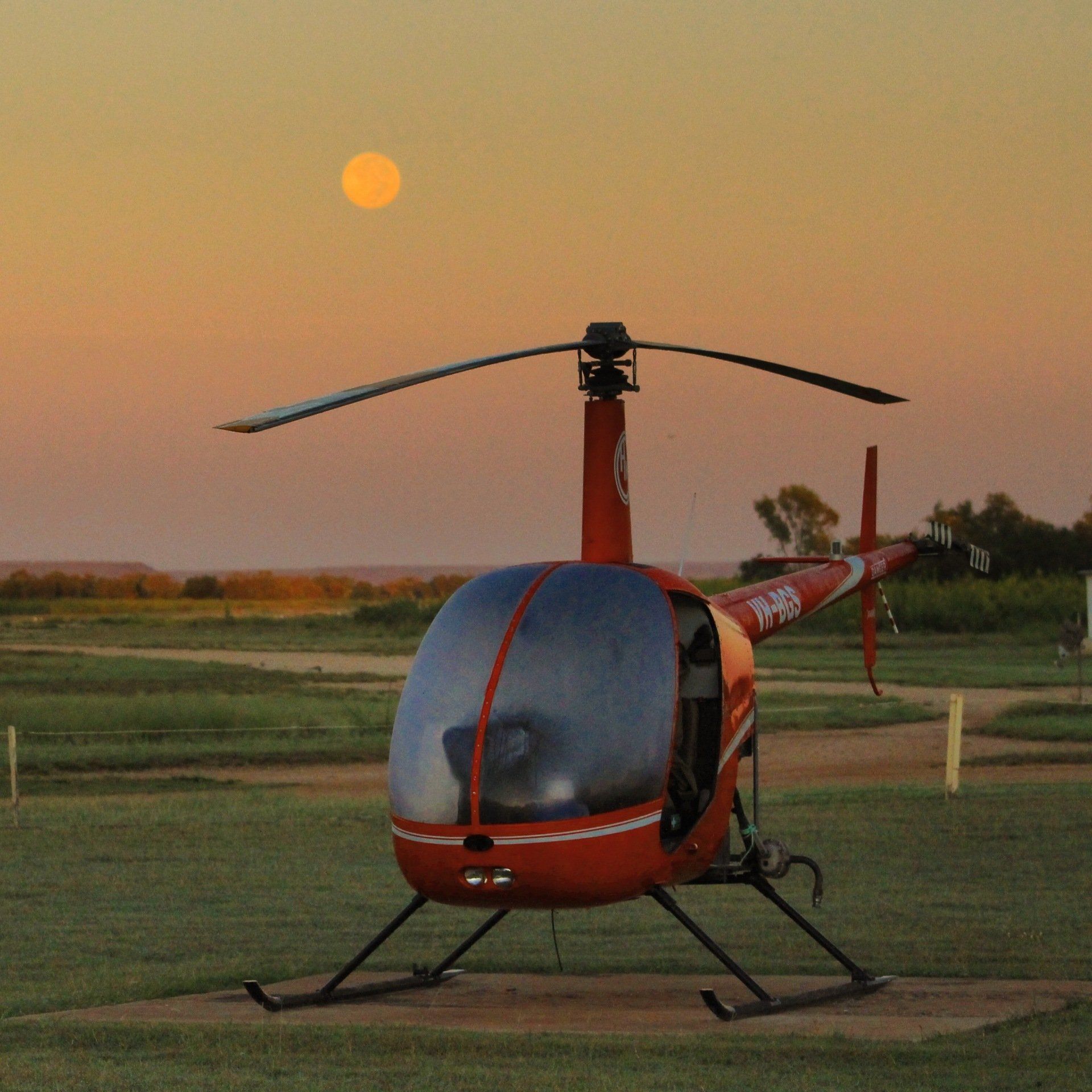 Red helicopter on a grassy field at sunset with the sun in the sky.