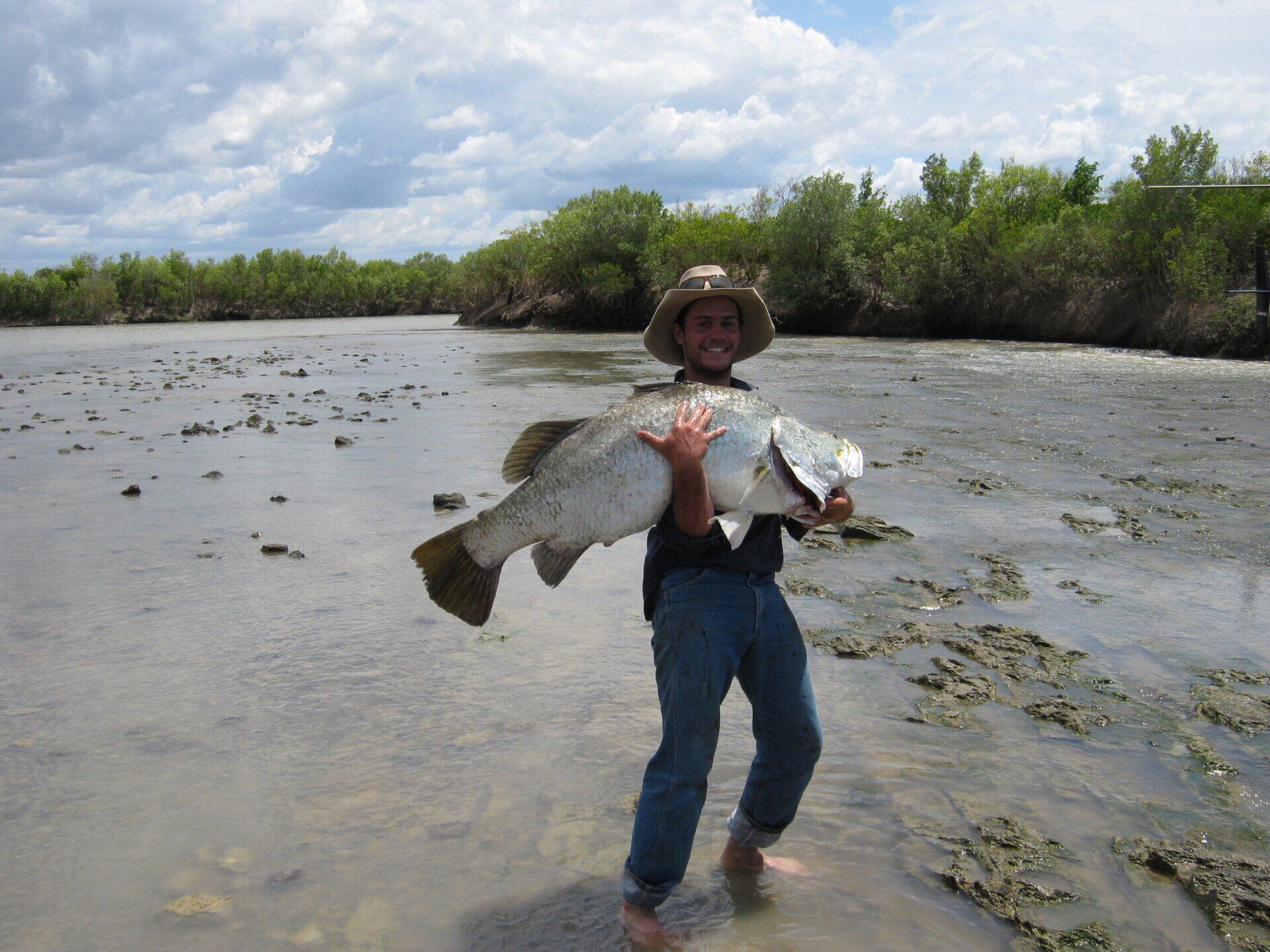 Man holding large silver barramundi fish, standing in muddy water, shoreline background.