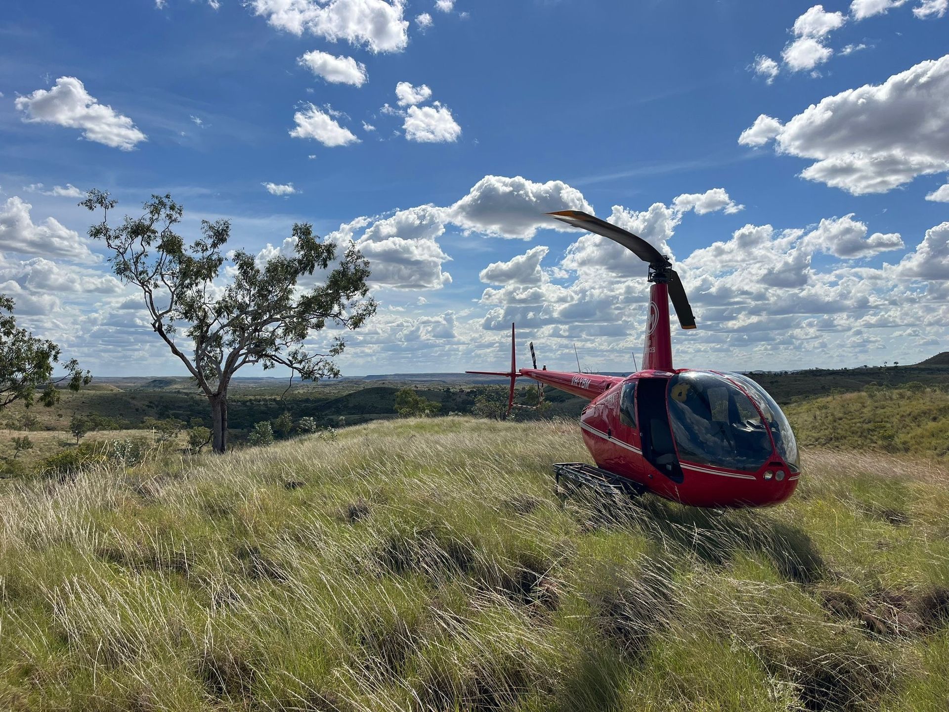 Red helicopter parked on a grassy hill under a partly cloudy sky.