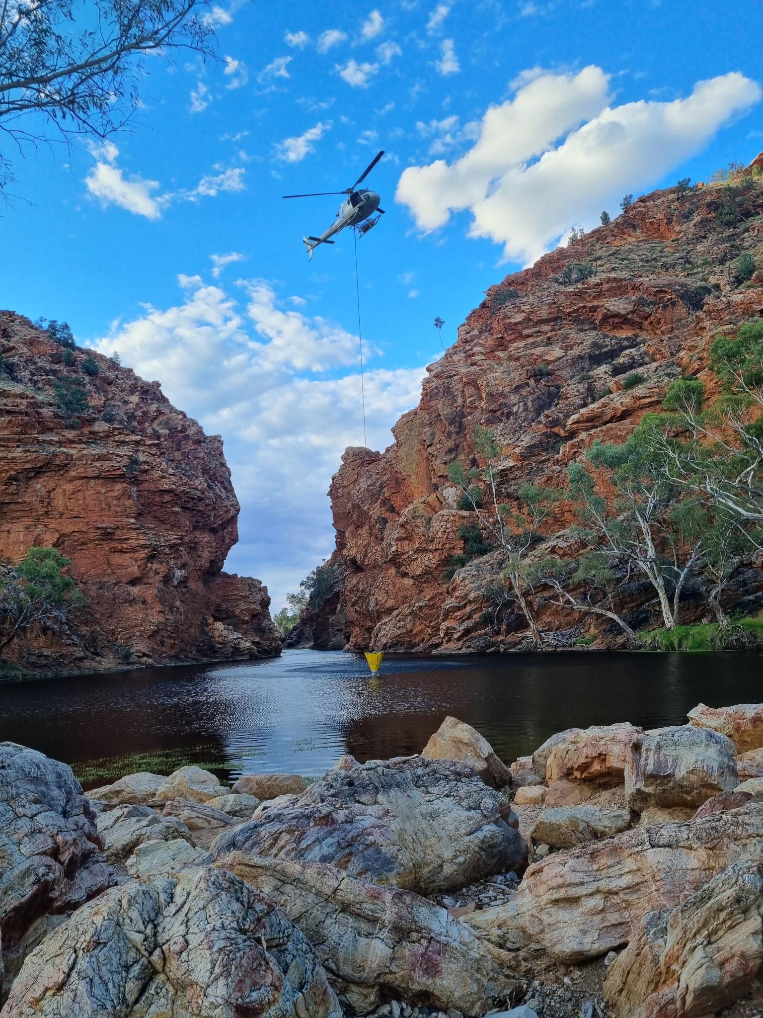 Helicopter over a canyon lake, hauling supplies. Red rock cliffs, blue sky with clouds.