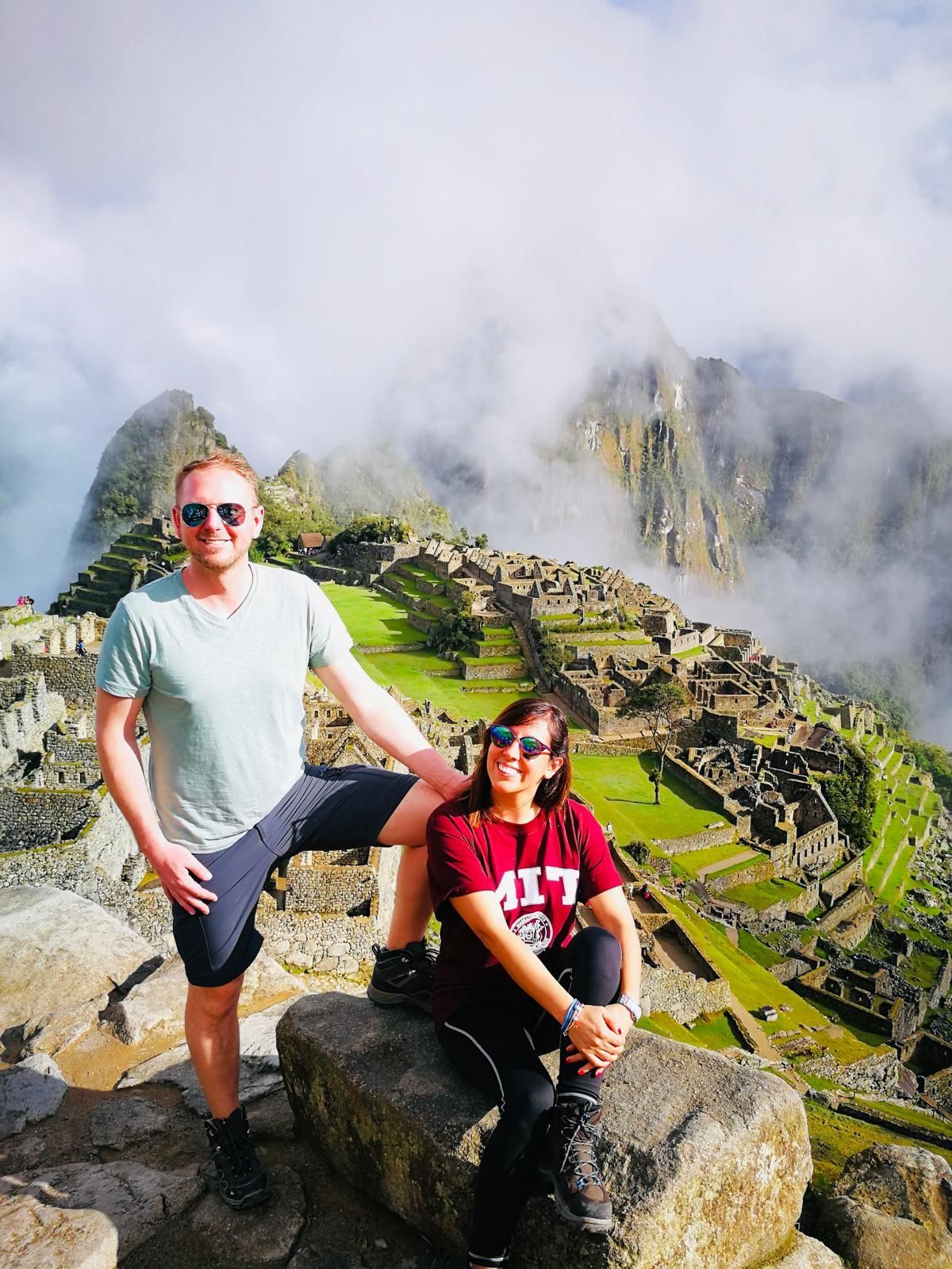 Pareja posa en Machu Picchu. Hombre con camisa azul, mujer con camisa roja. Ruinas y montaña al fondo.