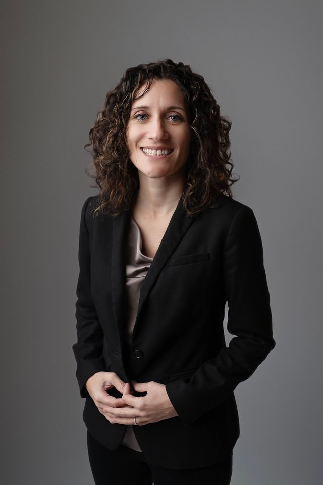 Woman with curly hair in a black blazer smiling. Gray background.