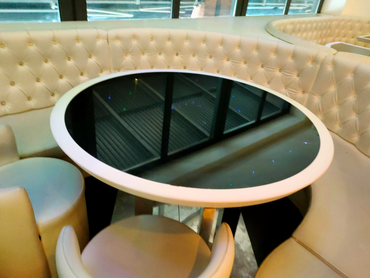 In un ristorante moderno, un tavolo circolare con piano nero è circondato da divanetti e sedie imbottiti di bianco.