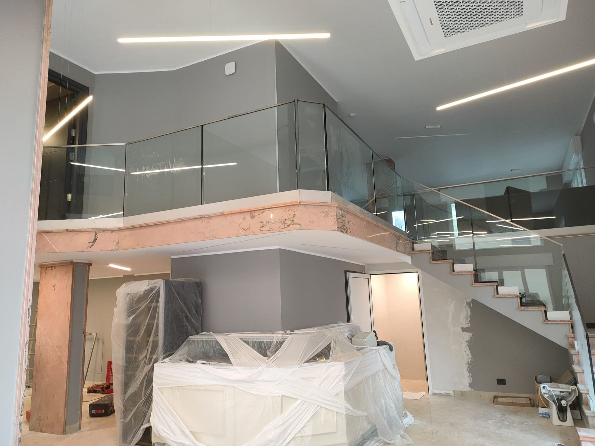 Vista interna di un moderno spazio a due piani in costruzione, con pareti grigie, ringhiere in vetro e oggetti rivestiti in plastica.