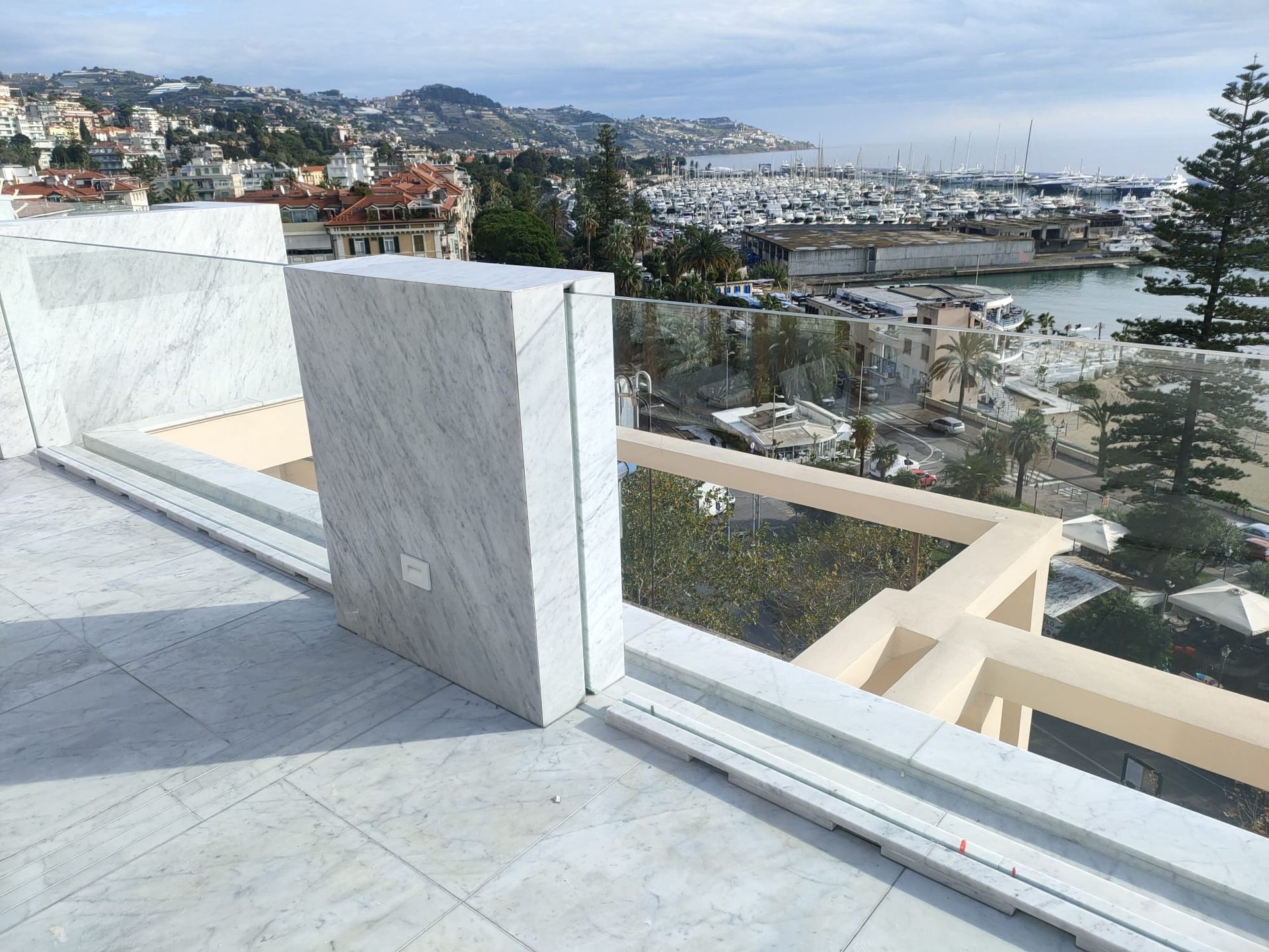 Una terrazza con balcone in marmo affacciata su una cittadina costiera, un porticciolo turistico e il mare, sotto un cielo luminoso e parzialmente nuvoloso.