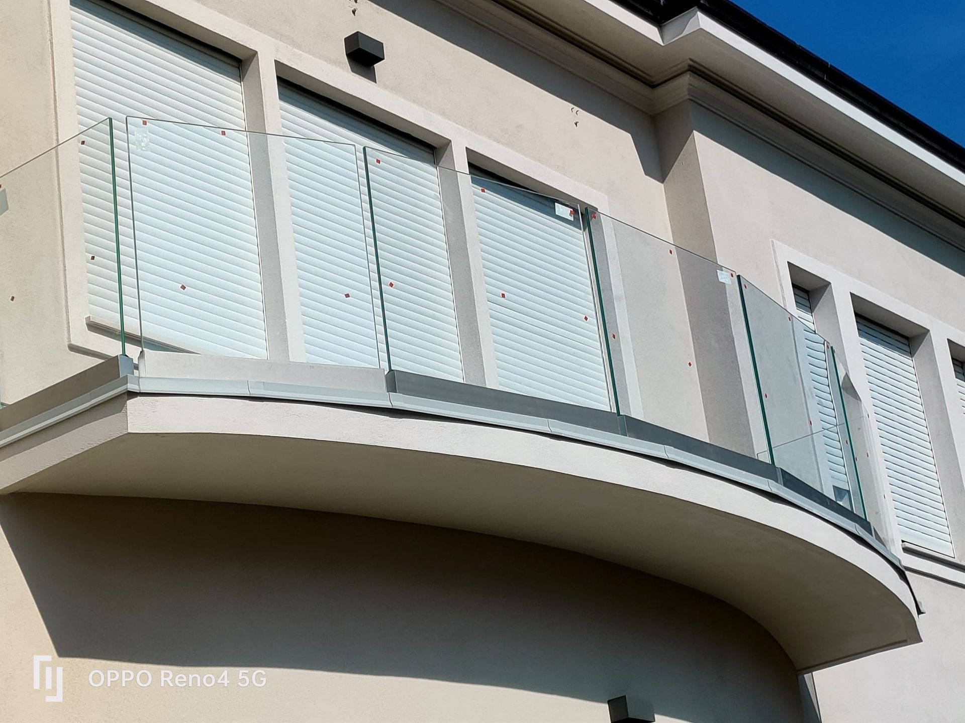 Un balcone residenziale curvo con ringhiera in vetro trasparente e finestre con persiane bianche su una parete esterna beige.