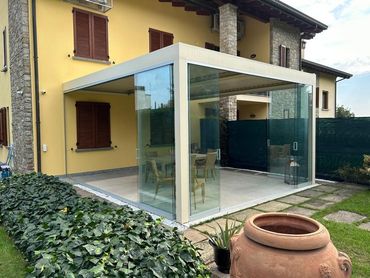 Una moderna pergola di colore chiaro con pareti in vetro, annessa a una casa gialla, con zona pranzo all'aperto.