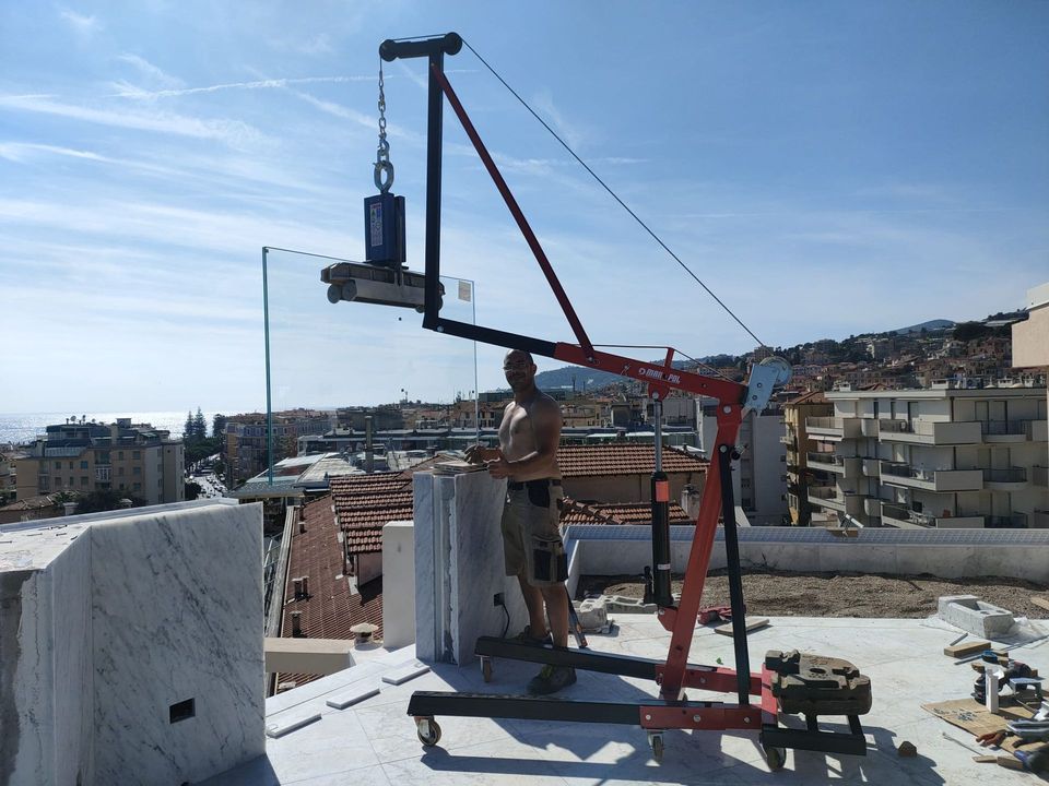 Una persona manovra una gru portatile rossa per sollevare una grande lastra di vetro su un tetto che si affaccia su una cittadina costiera.