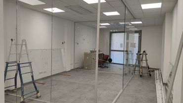 Vista interna di un ufficio non ancora ultimato, con pareti divisorie in vetro, due scale e materiali edili sul pavimento.