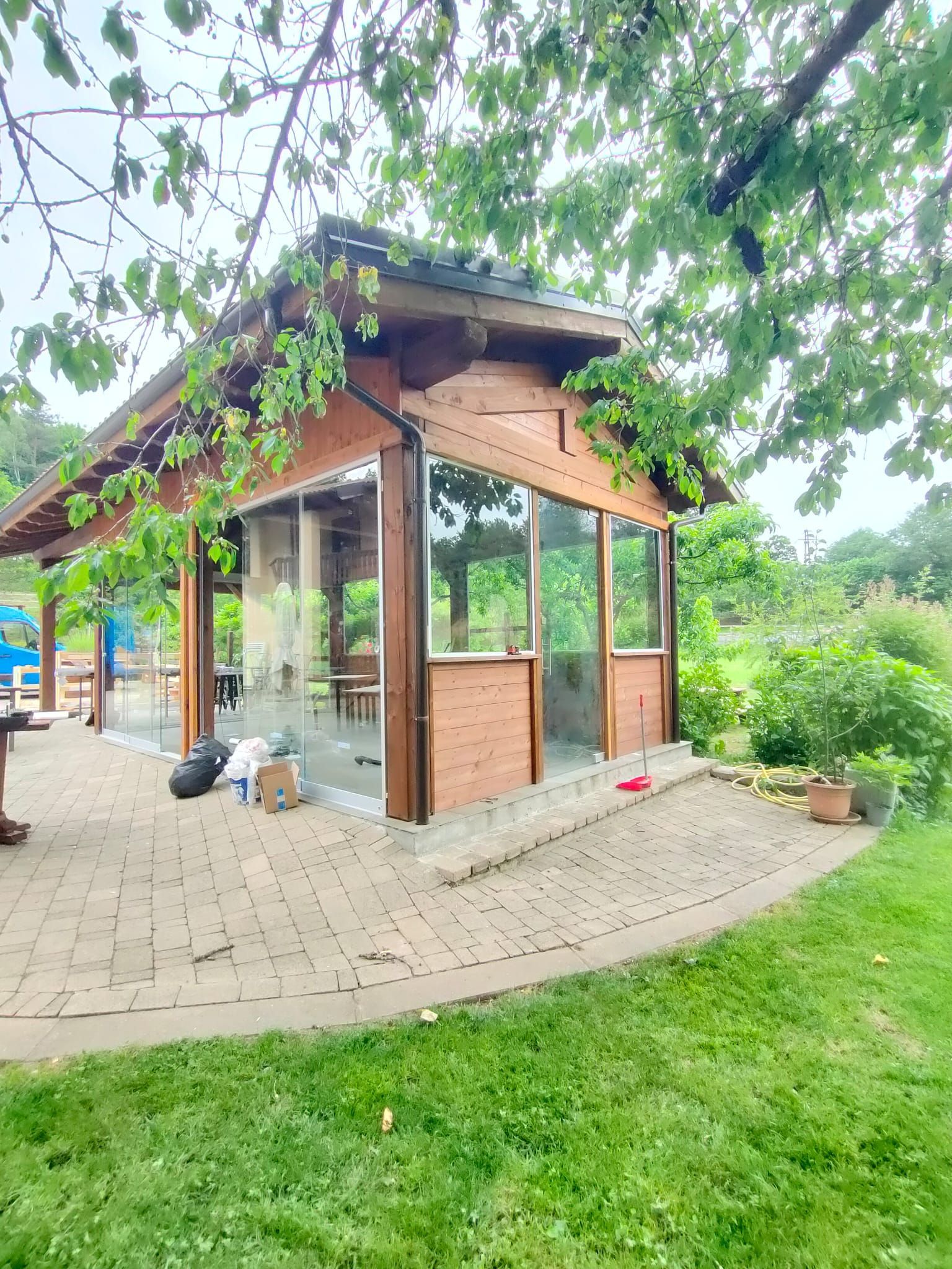 Un gazebo in legno con pareti in vetro e pavimento piastrellato, immerso in un rigoglioso giardino verdeggiante sotto i rami frondosi degli alberi.