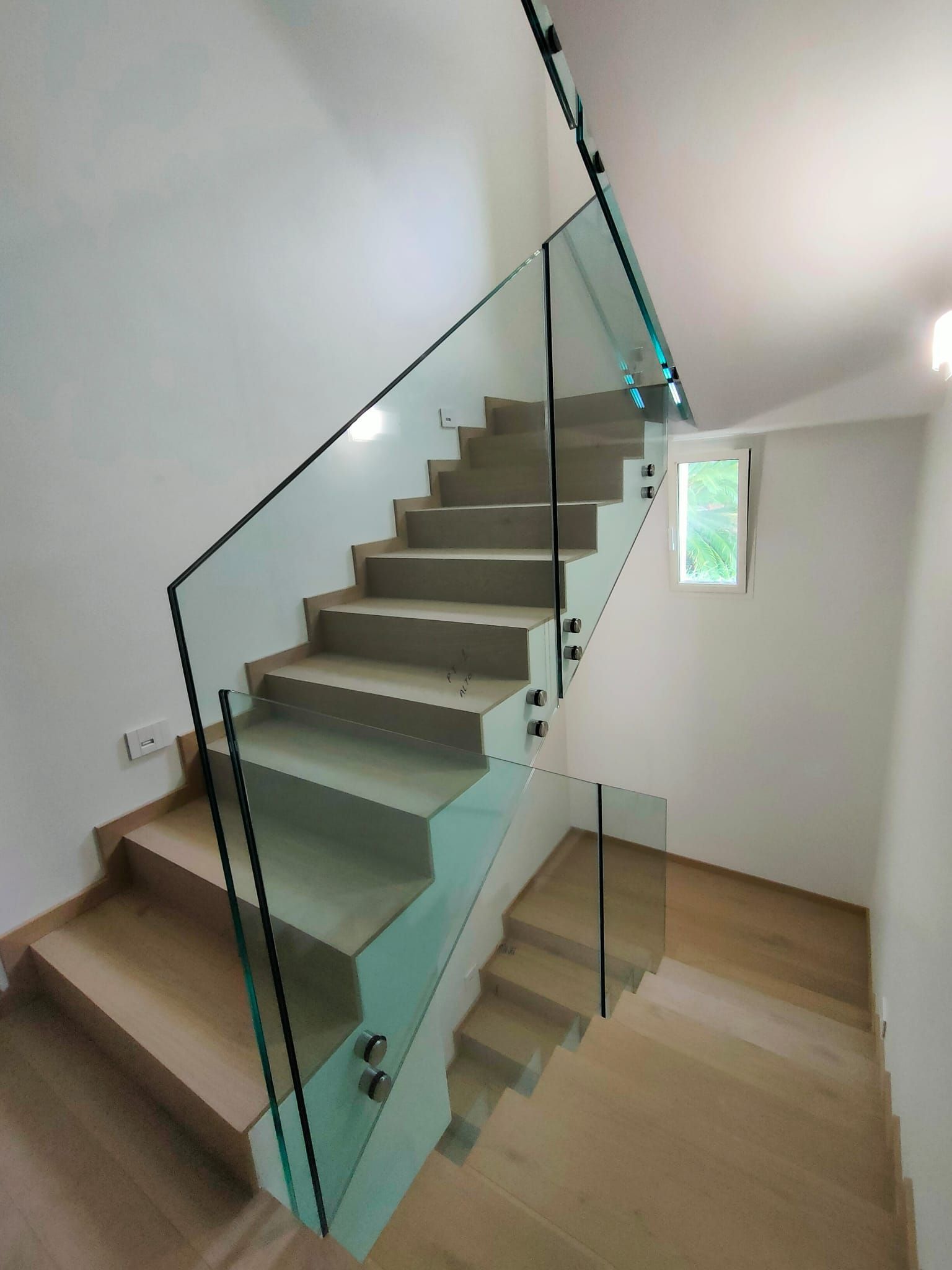 Una scala moderna con gradini in legno chiaro e ringhiera in vetro, che sale verso una piccola finestra in uno spazio luminoso e bianco.