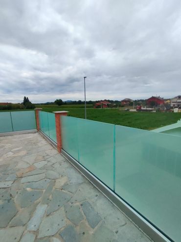 Un patio in pietra affacciato su un campo verde, con una ringhiera in vetro satinato e pilastri in mattoni sotto un cielo nuvoloso.