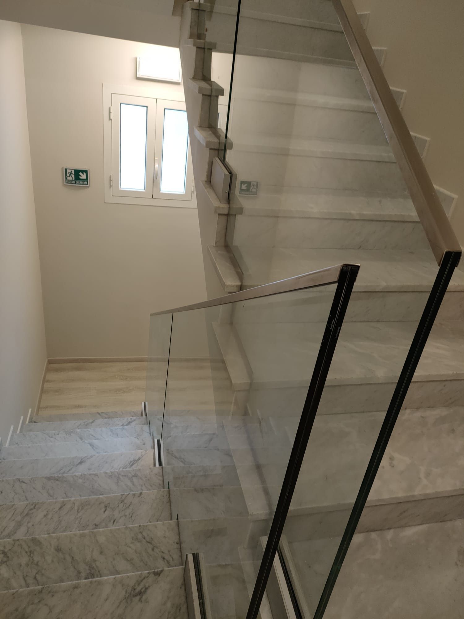 Una scala con gradini in marmo chiaro e ringhiere in vetro, vista dall'alto, che conduce a un piccolo pianerottolo con una finestra.