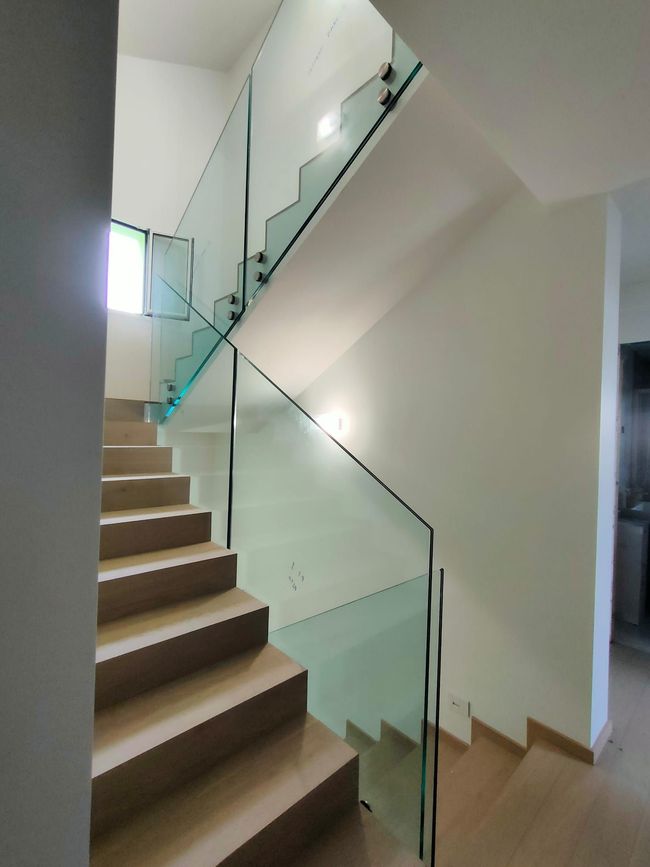 Una moderna scala interna con gradini in legno chiaro e una ringhiera in vetro trasparente che si estende fino al pianerottolo superiore.
