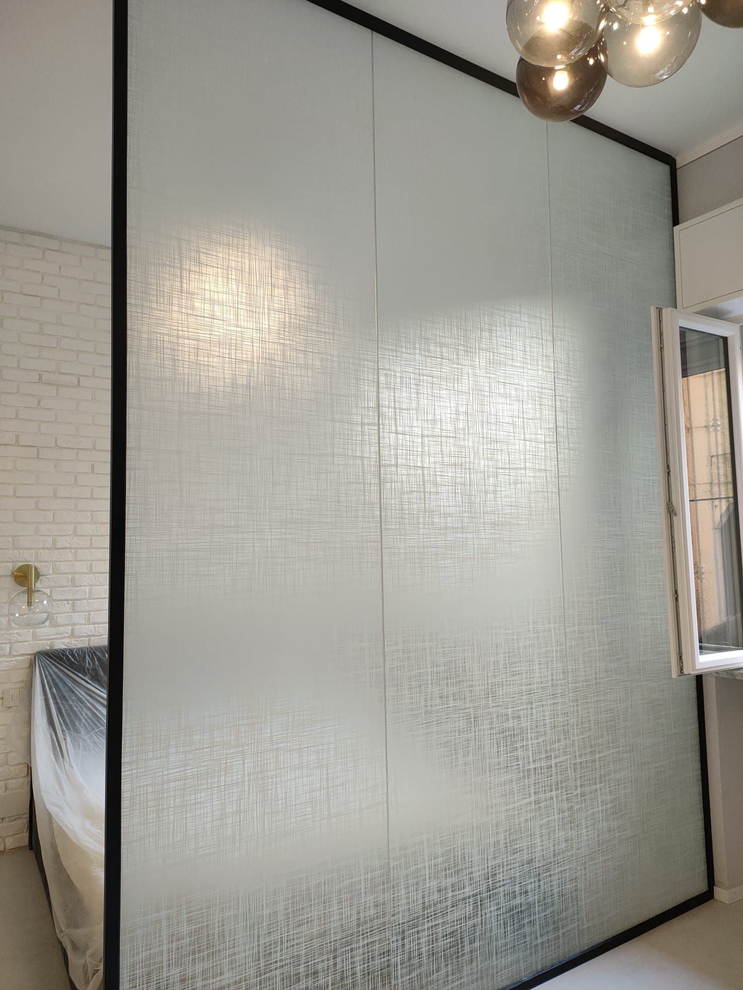 Una parete divisoria in vetro a tutta altezza con una trama satinata, inserita in una stanza con una parete in mattoni bianchi.