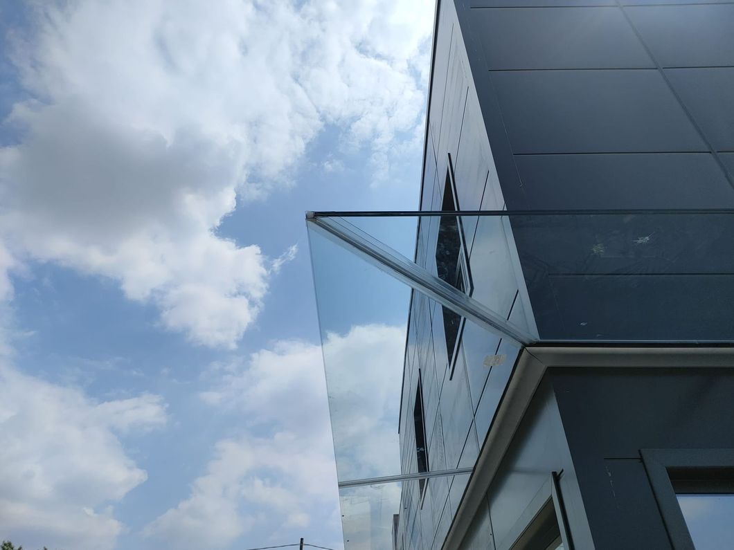 Una vista dal basso della facciata di un edificio moderno con una tettoia in vetro che si estende da un angolo, sullo sfondo di un cielo luminoso e nuvoloso.