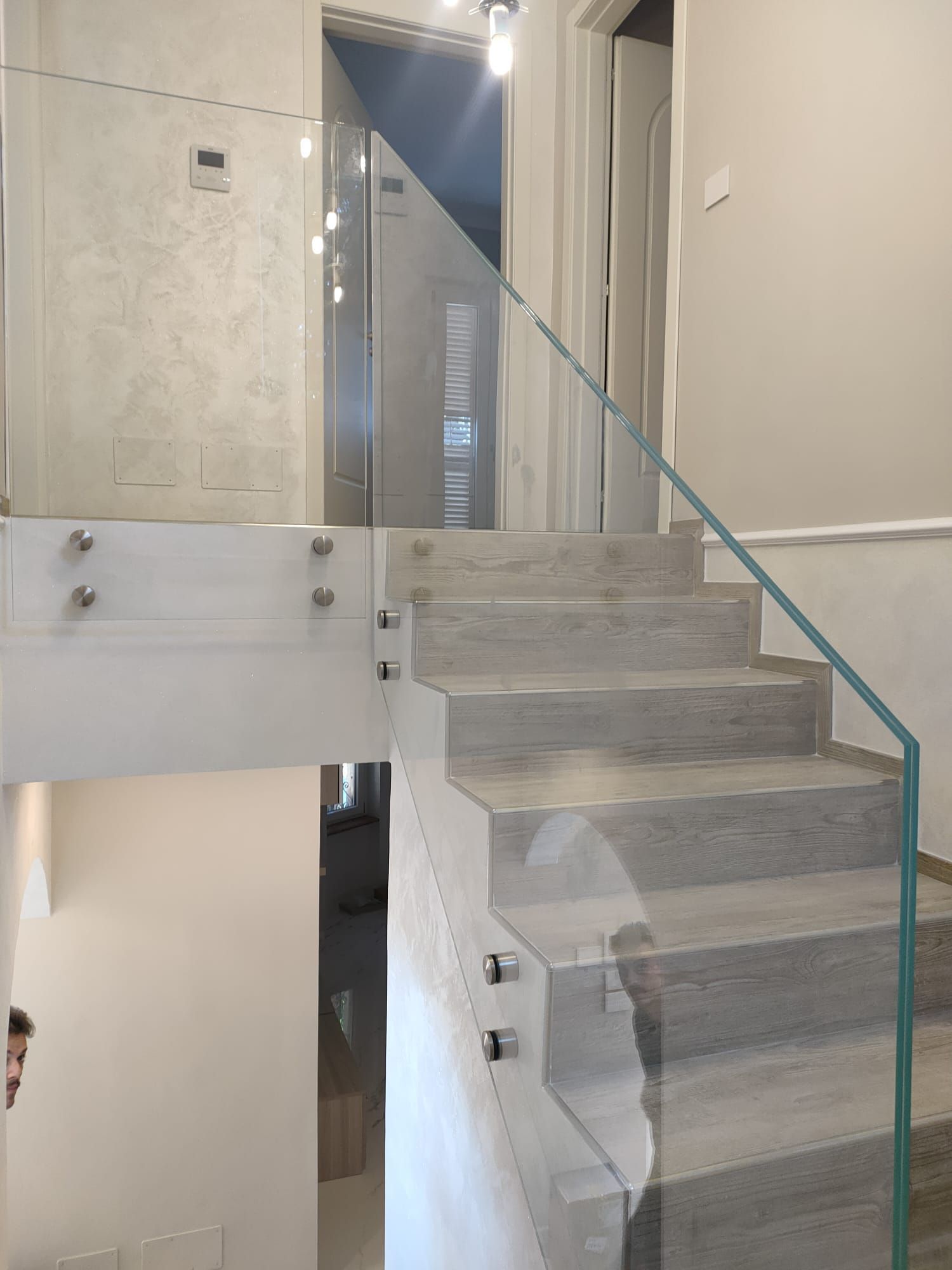 Una scala con gradini in legno chiaro e una moderna ringhiera in vetro senza telaio, fissata con distanziatori metallici.