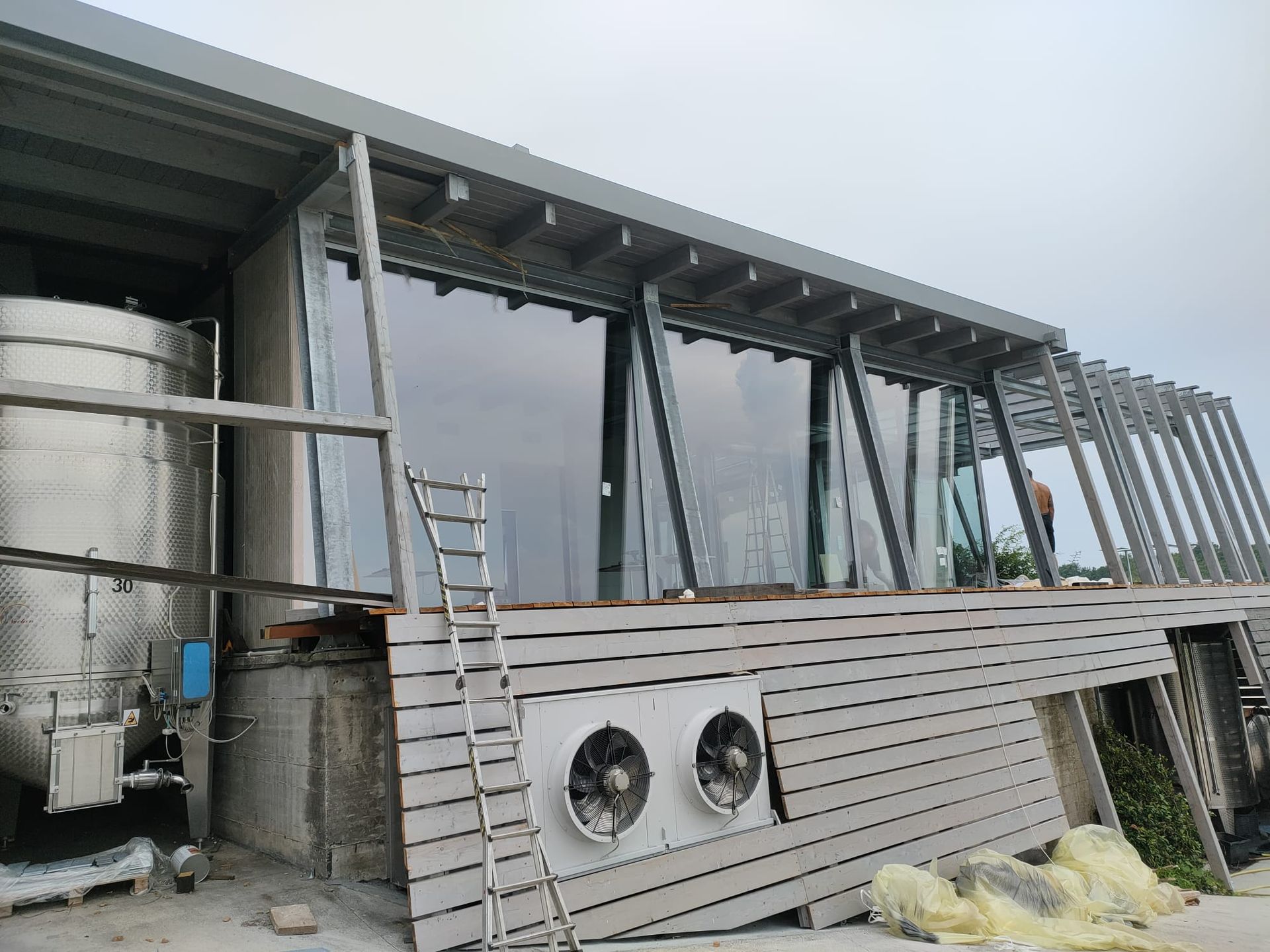 Un moderno edificio in costruzione presenta ampie vetrate, rivestimenti in legno, un serbatoio metallico e ventilatori esterni per il raffreddamento.