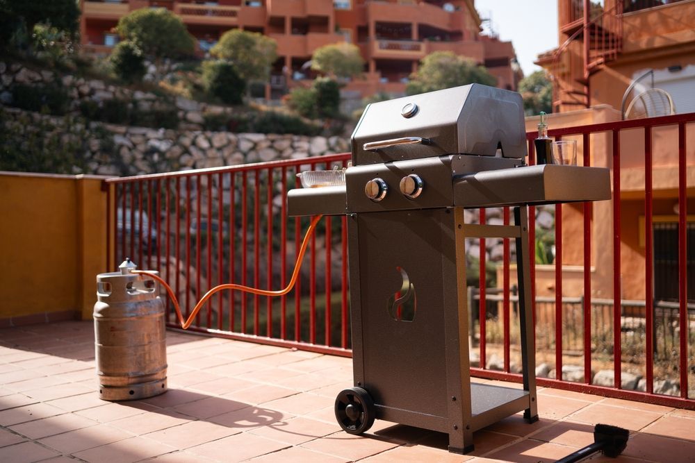 Gas grill with propane tank on a patio, red railing, and building in the background.
