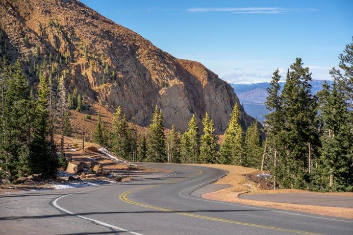 Curving mountain road with evergreen trees, brown rocky hillside, and clear blue sky.