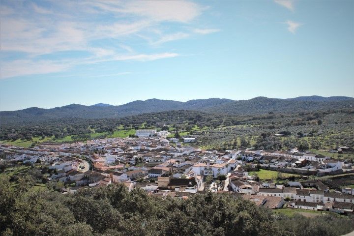 A small, white-walled town nestled in a valley surrounded by rolling green hills and olive groves under a blue sky.
