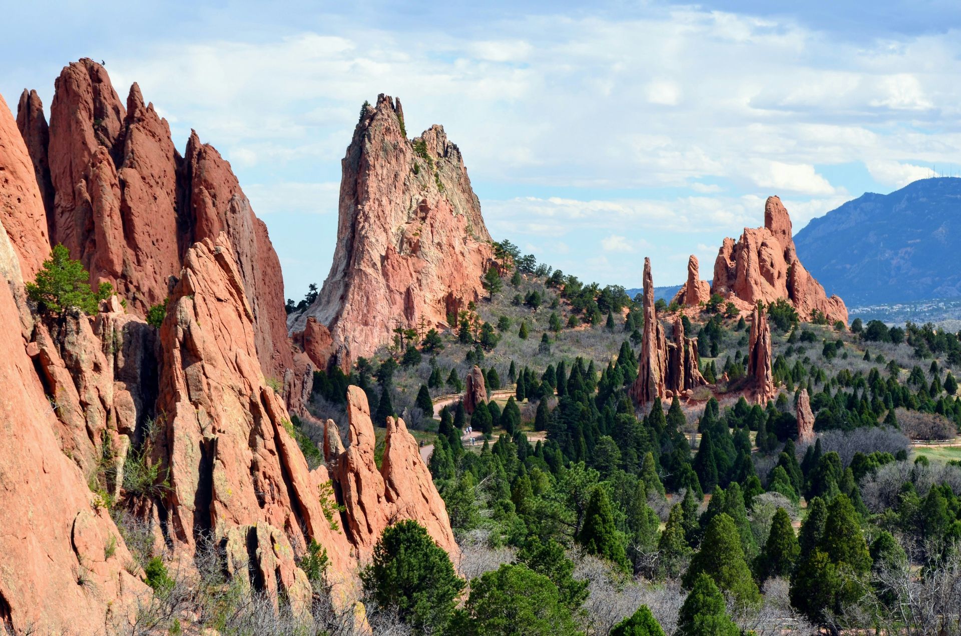 Jagged red sandstone rock formations rise above a forest of green pine trees under a cloudy blue sky.