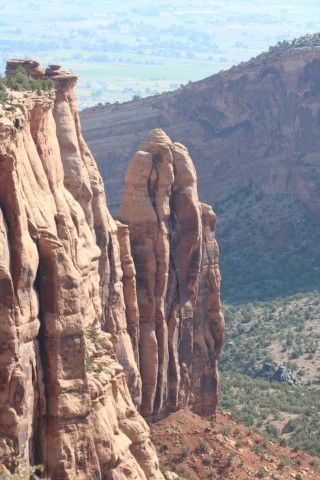 A tall, slender red rock monolith stands separated from a nearby canyon wall, overlooking a sparse, vegetated valley.