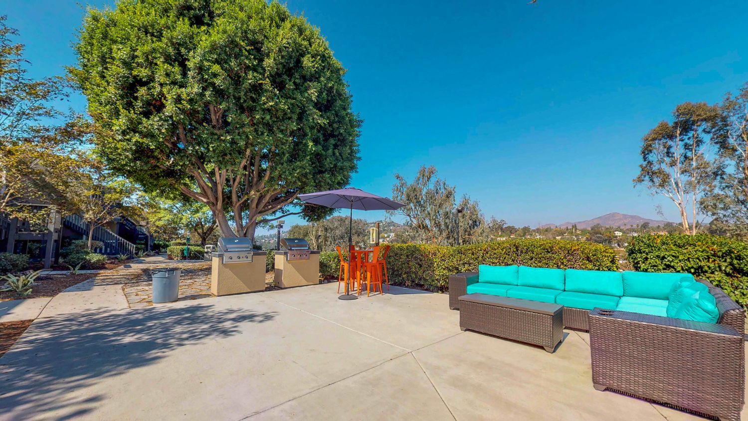 A sunny outdoor patio features a large turquoise sectional sofa, a small dining set with an umbrella, and two grills.