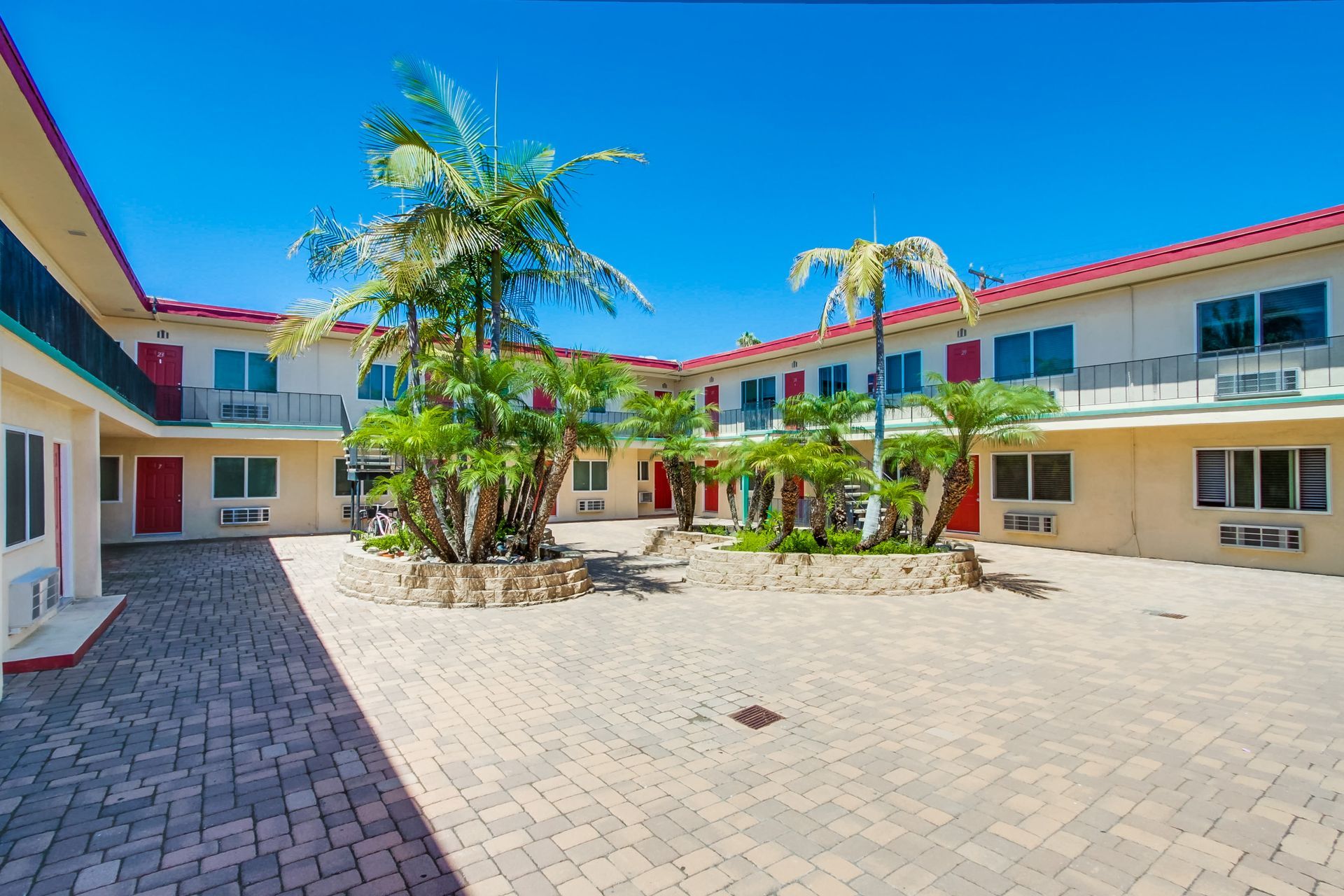 An outdoor courtyard of a hotel featuring a brick-paved ground, lush green palm trees, and two-story cream-colored rooms.