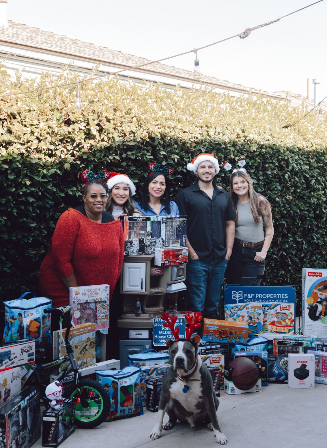 Five people and a dog wearing reindeer antlers sit surrounded by many boxed toys in front of a green hedge.