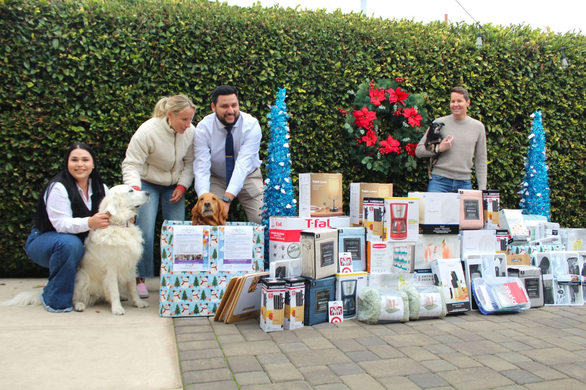 Three people and three dogs pose in front of a hedge decorated with a wreath and wrapped gifts for a holiday event.