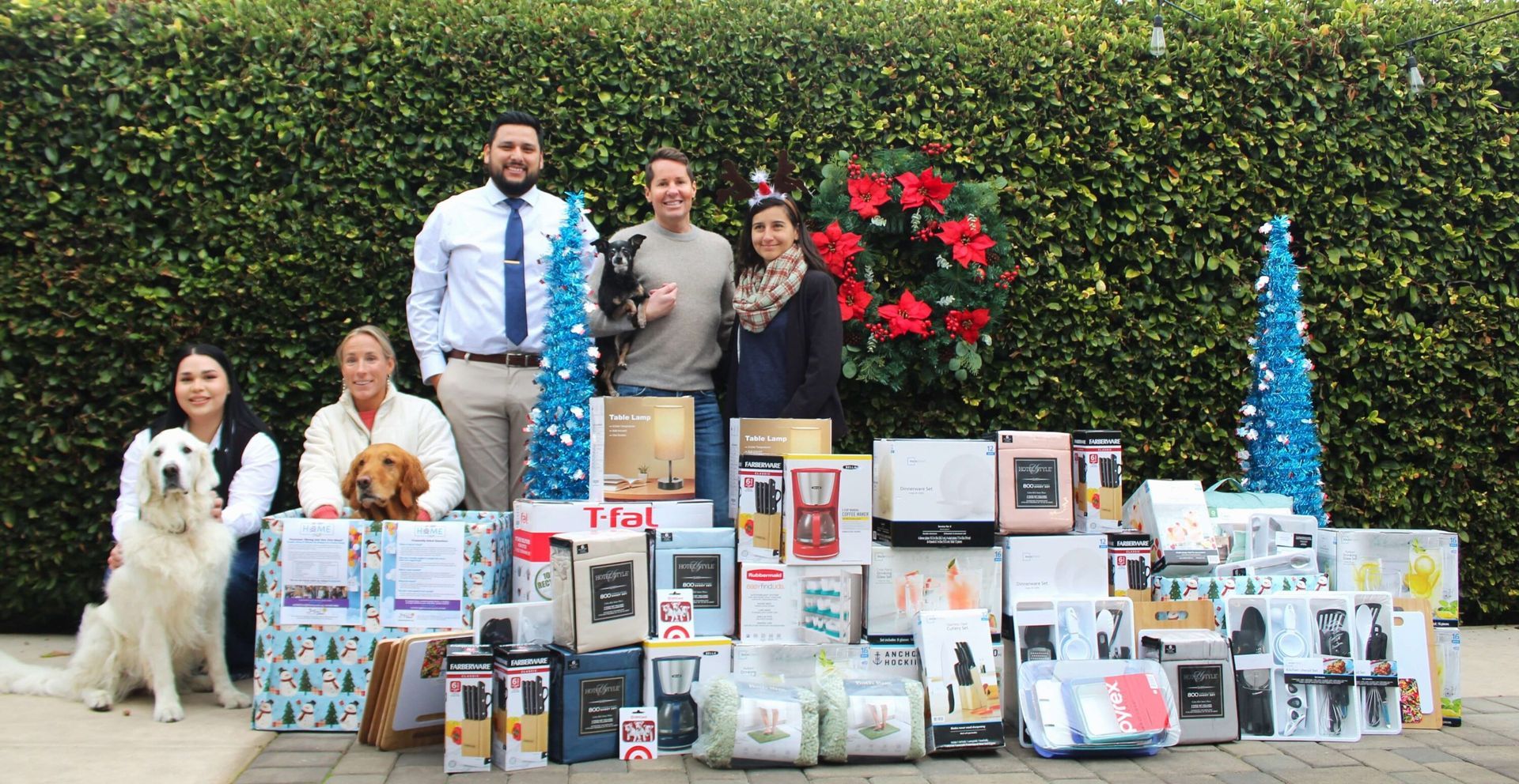 A group of four people and two dogs pose with many wrapped holiday gifts in front of a hedge with Christmas decorations.