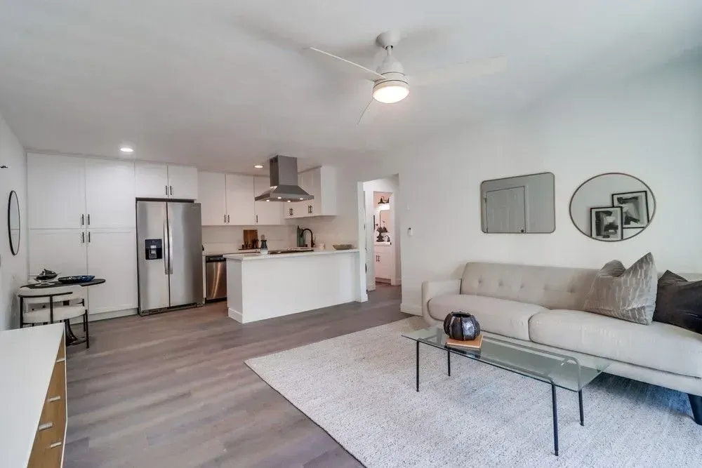 A bright, open-concept room with light wood floors, a white sofa, glass coffee table, and an adjacent modern kitchen.