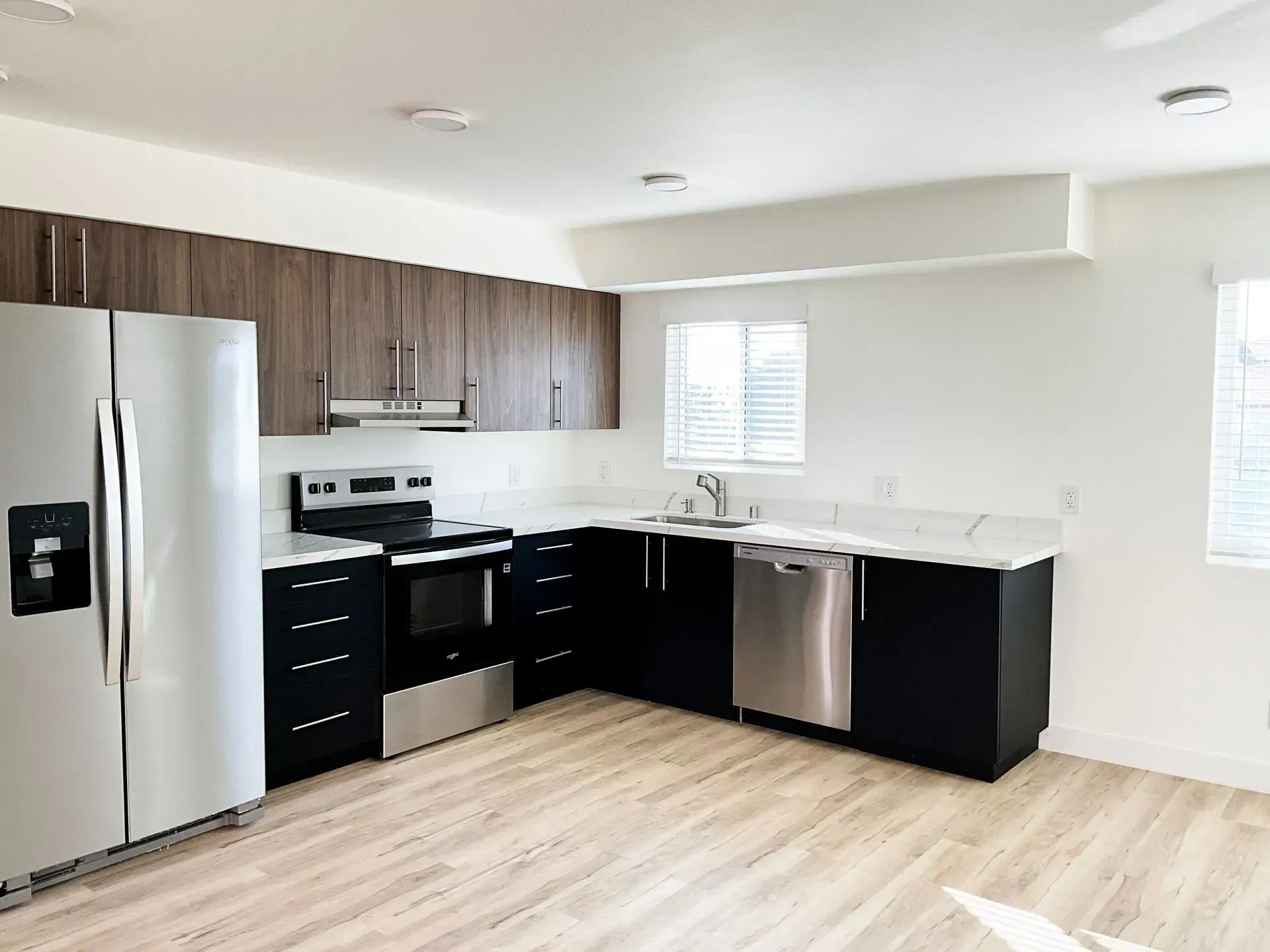 A modern kitchen with light wood-look floors, brown upper cabinets, black lower cabinets, and stainless steel appliances.