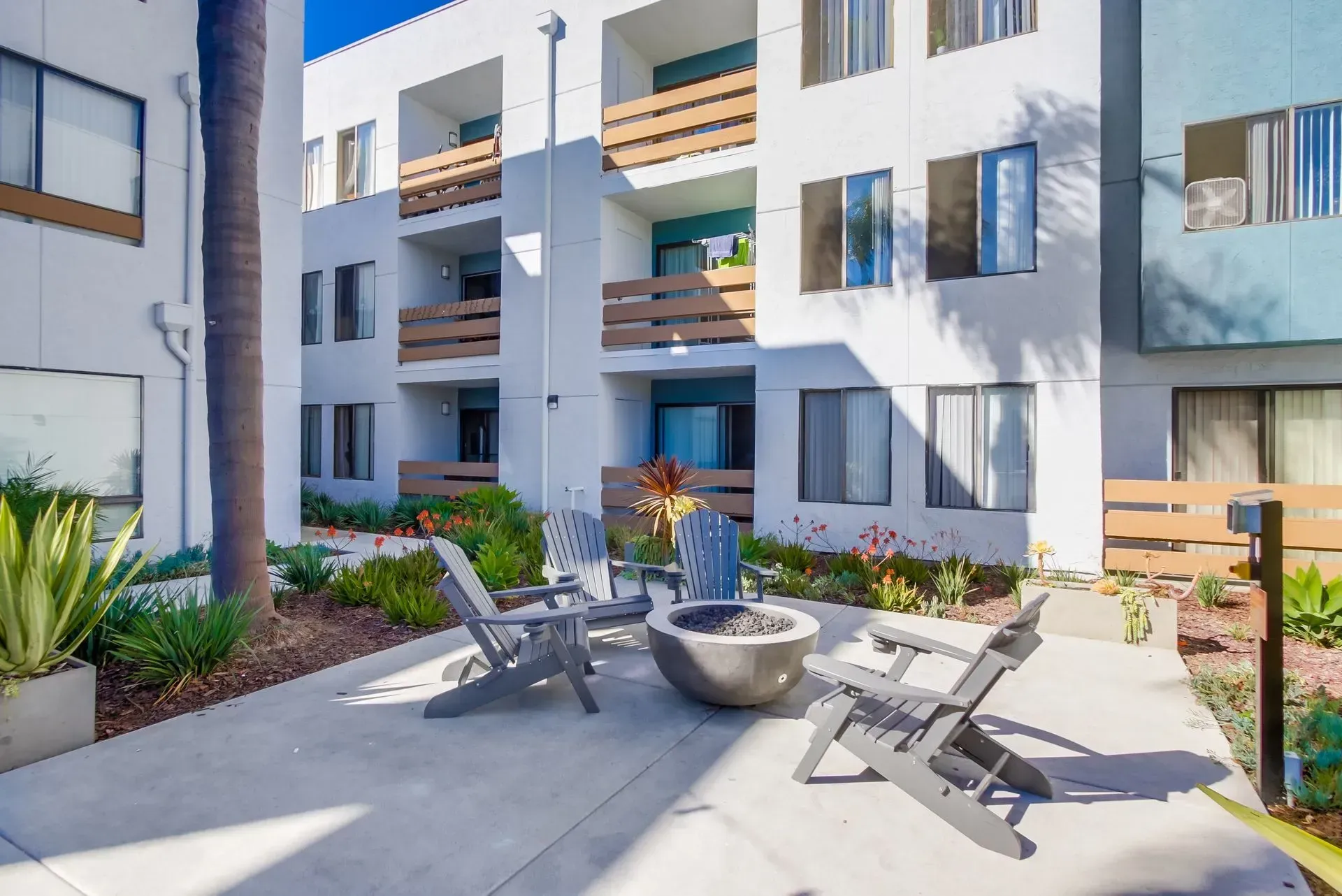 A patio with gray adirondack chairs surrounding a round fire pit outside a multi-story apartment building.