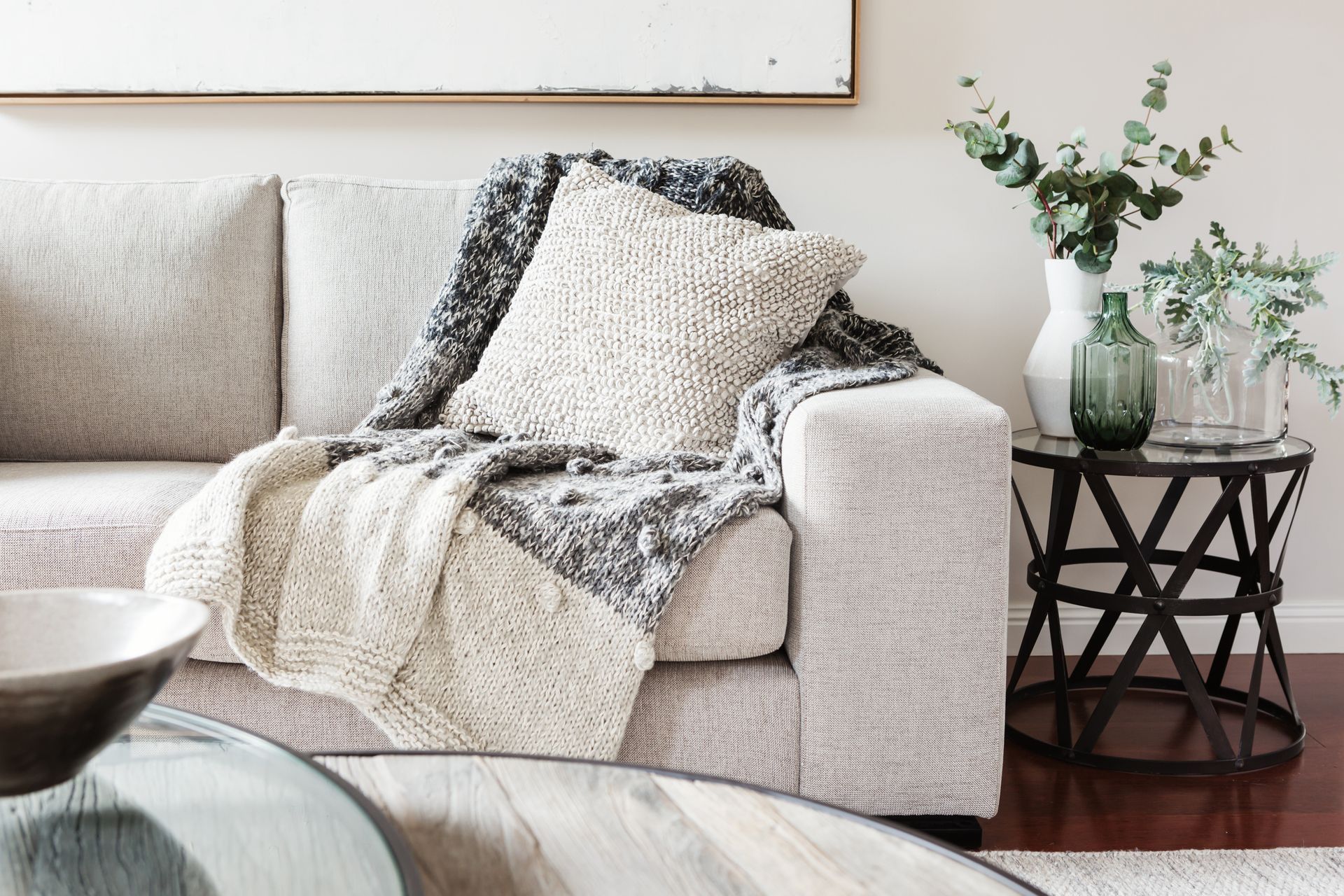 A beige couch with a knit blanket and pillow sits beside a small black side table holding vases and greenery.